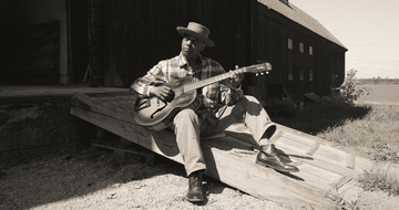 A black man wearing a brimmed hat, plaid shirt, straight leg trousers and smart shoes, sits outside on a wooden ramp and strums his guitar