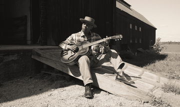 A black man wearing a brimmed hat, plaid shirt, straight leg trousers and smart shoes, sits outside on a wooden ramp and strums his guitar