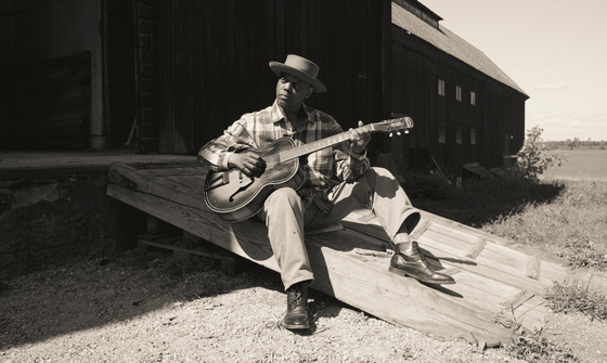 A black man wearing a brimmed hat, plaid shirt, straight leg trousers and smart shoes, sits outside on a wooden ramp and strums his guitar