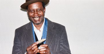 Errol, a black man with a brown fedora and a grey blazer smiles as he holds a harmonica