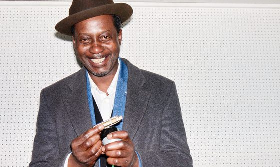 Errol, a black man with a brown fedora and a grey blazer smiles as he holds a harmonica