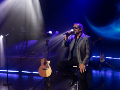A man in a leather jacket and sunglasses sits on a stool as he sings into a handheld microphone