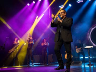 A man in a black suit and sunglasses claps on stage surrounded by an array of musicians