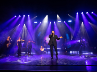 Lit by blue lighting a man in a black suit sings on stage alongside three musicians