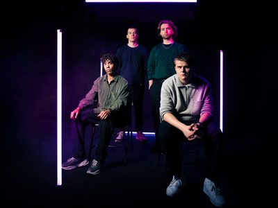 Two men sit down and two men stand up behind them, all are dressed in casual clothing and lit by three strip bars of lights