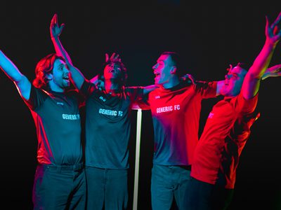 Four young men in red football shirts stand with their arms raised up or resting across each other's shoulders. All of them cheer in unison.