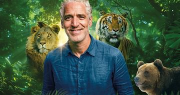Gordon Buchanan, a smiling man in a blue shirt standing in a lush jungle with a lion, tiger, and bear in the background. Sunlight filters through dense foliage