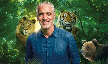 Gordon Buchanan, a smiling man in a blue shirt standing in a lush jungle with a lion, tiger, and bear in the background. Sunlight filters through dense foliage