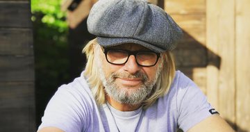 A photo of Henry Cole, an older white man with long blonde hair, a short neat white beard, glasses, and grey baker boy hat stood outside of a shed