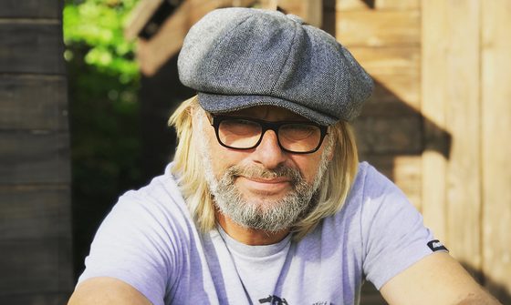 A photo of Henry Cole, an older white man with long blonde hair, a short neat white beard, glasses, and grey baker boy hat stood outside of a shed