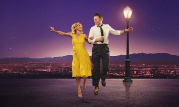 A woman in a yellow dress holding the arm of a man in a white shirt with black tie and trousers as they look at each other dancing, there is a lamppost behind them and a city in the distance. The photo is a homage to La La Land