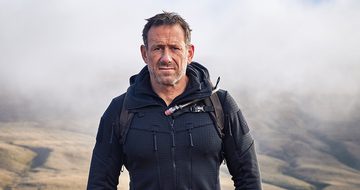 A photo of Jason Fox walking outside with fog behind him in a jacket with a backpack. He has a serious expression
