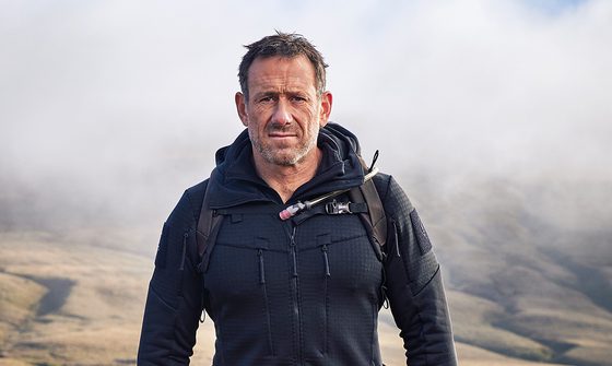 A photo of Jason Fox walking outside with fog behind him in a jacket with a backpack. He has a serious expression