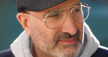 A man with grey stubble, gold rimmed glasses and a grey flat cap