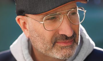 A man with grey stubble, gold rimmed glasses and a grey flat cap