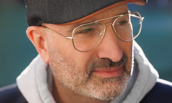 A man with grey stubble, gold rimmed glasses and a grey flat cap