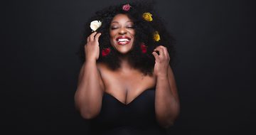 Judi Love on a black background in a black dress grinning while touching the white, red, and yellow flowers in her hair