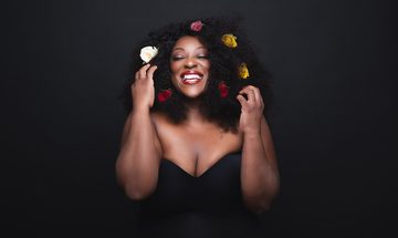 Judi Love on a black background in a black dress grinning while touching the white, red, and yellow flowers in her hair