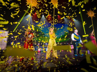 A photo of a man in red, a woman in yellow, and a woman in pink all holding up sun drawings on sticks with Justin the middle. The photo is obscured by a large amount of yellow confetti falling down