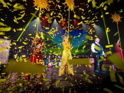 A photo of a man in red, a woman in yellow, and a woman in pink all holding up sun drawings on sticks with Justin the middle. The photo is obscured by a large amount of yellow confetti falling down