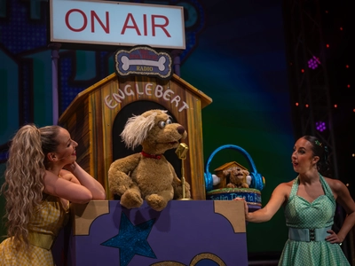 The woman in a yellow and green dress are stood either side of a dog puppet who has a gold microphone and is in front of a dog house that reads 'Englebert' with a large On Air sign above