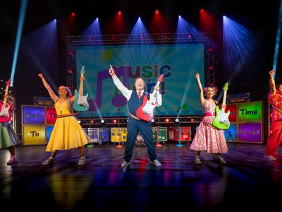 Five people on stage all holding different coloured electric guitars in their left hand with right in the air. Left to right, woman in a green dress, woman in a yellow dress, Justin Fletcher, woman in a white and pink dress, man in red trousers and jacket