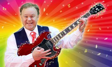Justin Fletcher in a white shirt, navy waistcoat, and red tie with white polka dots holding a red electric guitar and smiling in front of a multi-coloured background with golden music notes and stars