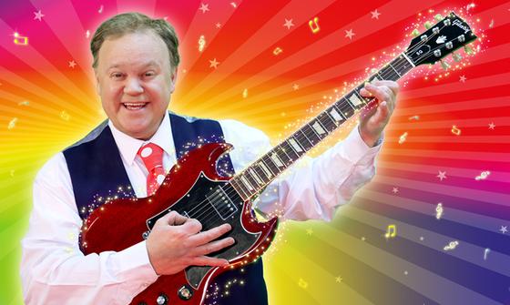 Justin Fletcher in a white shirt, navy waistcoat, and red tie with white polka dots holding a red electric guitar and smiling in front of a multi-coloured background with golden music notes and stars