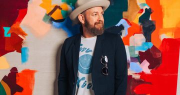 Kristian Bush, stands against a multicoloured wall. He looks to his side as he leans against the wall. He's wearing a beige fedora, a navy blazer with sunglasses hanging from the breast pocket, and a white t-shirt with a blue smiley face on it