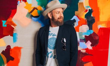 Kristian Bush, stands against a multicoloured wall. He looks to his side as he leans against the wall. He's wearing a beige fedora, a navy blazer with sunglasses hanging from the breast pocket, and a white t-shirt with a blue smiley face on it