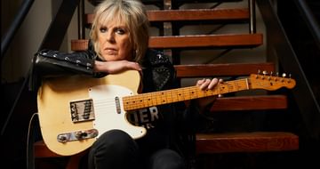 An older woman sat on some stairs in a studded leather jacket leaning on an electric guitar on her lap