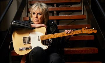 An older woman sat on some stairs in a studded leather jacket leaning on an electric guitar on her lap