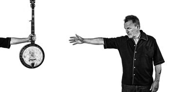 Martin Simpson wears a black denim shirt and jeans, he reaches his right arm out reaching for a banjo which is held up by an arm across the other side of the image