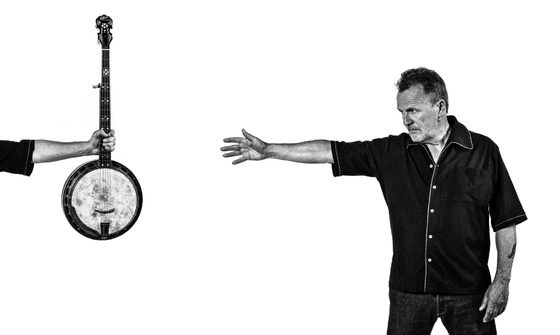 Martin Simpson wears a black denim shirt and jeans, he reaches his right arm out reaching for a banjo which is held up by an arm across the other side of the image