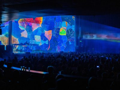 A view of the whole auditorium watching the show, a blue, red and yellow abstract design is projected on stage