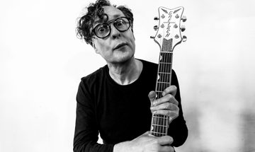 A black and white photo of Miles Hunt in a black long sleeve shirt holding a guitar by it's neck