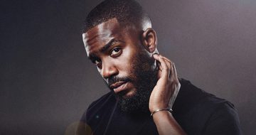 Mo Gilligan in a black tshirt on a black background with a small amount of light on his face