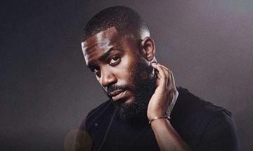 Mo Gilligan in a black tshirt on a black background with a small amount of light on his face