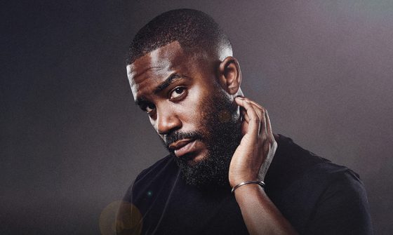 Mo Gilligan in a black tshirt on a black background with a small amount of light on his face