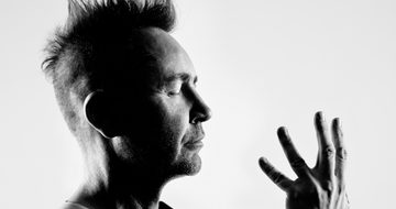 a black and white photo of Nigel Kennedy looking side on from shoulders upwards. He has his eyes closed and is looking towards one hand which is held up