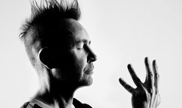 a black and white photo of Nigel Kennedy looking side on from shoulders upwards. He has his eyes closed and is looking towards one hand which is held up