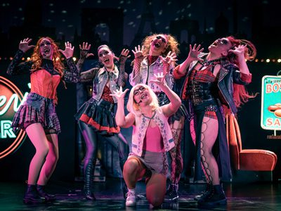 A vibrant stage performance with five actors in eclectic costumes, striking dynamic poses, with bright lights and neon signs in the background
