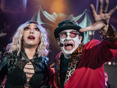Two performers on stage in dramatic attire. A woman with blonde hair and leather outfit, and a man in a vibrant costume, both with intense expressions