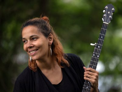Rhiannon smiles as she looks to the side of her. She's wearing all black and is holding the neck of a banjo