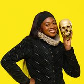 Efé Agwele, a black woman with long straight hair dressed in all black Tudor style clothes, with a white neck ruffle. She is smiling and holding up a skull.