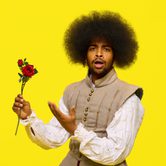 A black man with a large afro and short beard and moustache wearing a white shirt with a Tudor style beige tunic, red velvet trousers, and black trainers. He is holding up a rose looking surprised