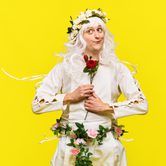 A white man with a long white wig in a long sleeve white dress and blue trainers, with flowers on his head, around his waist, and in his hands seemingly floating in the air