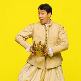 An Asian man with short black hair in a long sleeve beige tunic, with matching puffy trousers, and black trainers holding a crown, while dancing and smiling