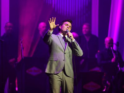 A man, lit in purple light and wearing a grey suit, sings as he stretches his hand out in front of him