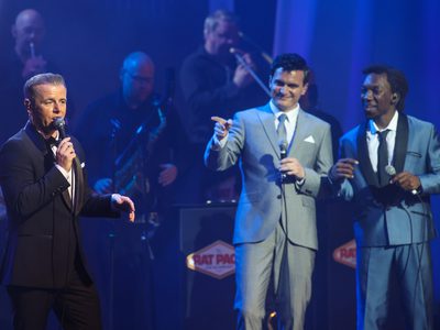 Three men acting as Frank Sinatra, Sammy Davis Jr and Dean Martin all stand on stage lit in blue light. One of them sings while the other two watch and point at him